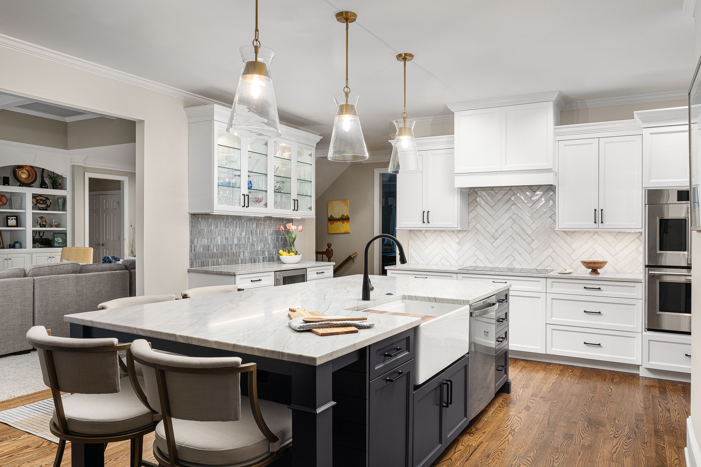 A white kitchen with a dark gray island, featuring a farmhouse sink, three pendant lights, and stainless steel appliances.