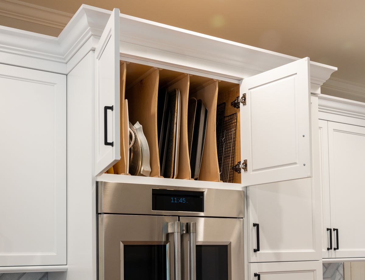 A kitchen features white cabinets, a stainless steel double oven, and an upper cabinet with open doors revealing vertical storage for baking sheets.