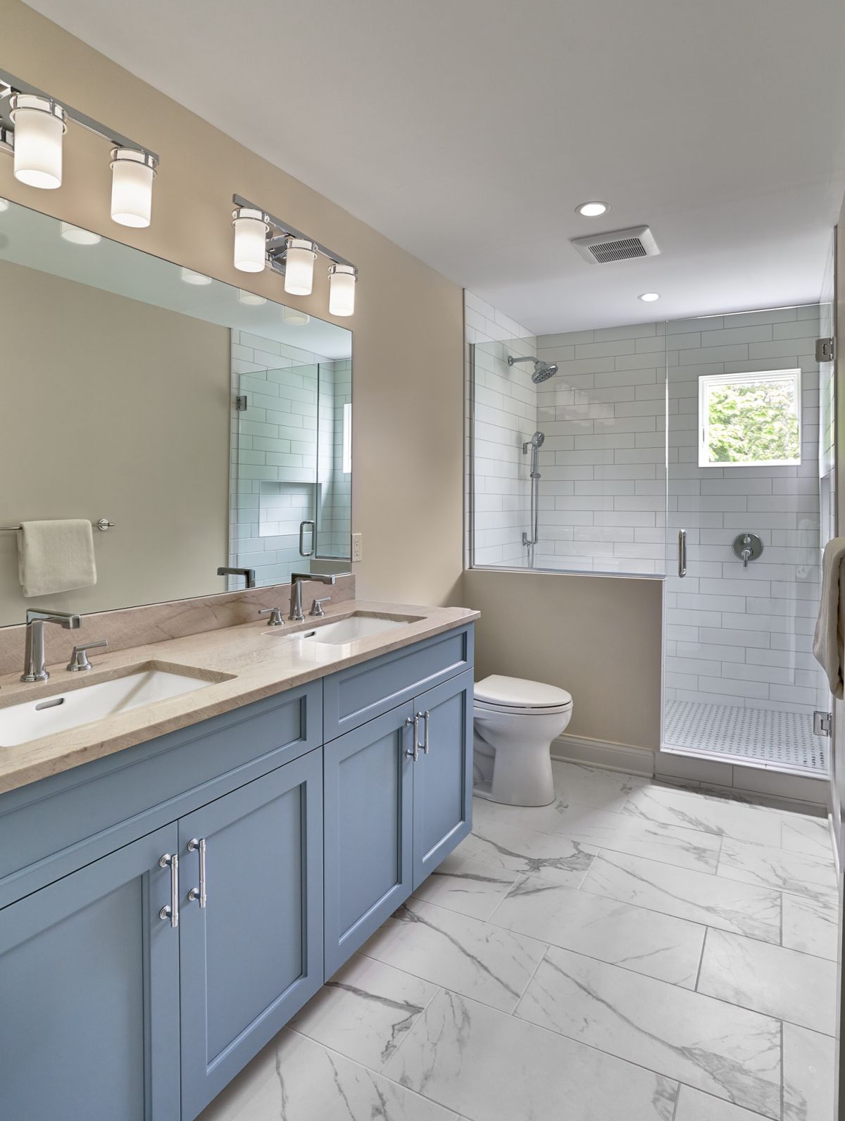 A bathroom with a light blue double vanity, a toilet, a walk-in shower with subway tiles, and a large mirror with vanity lights.