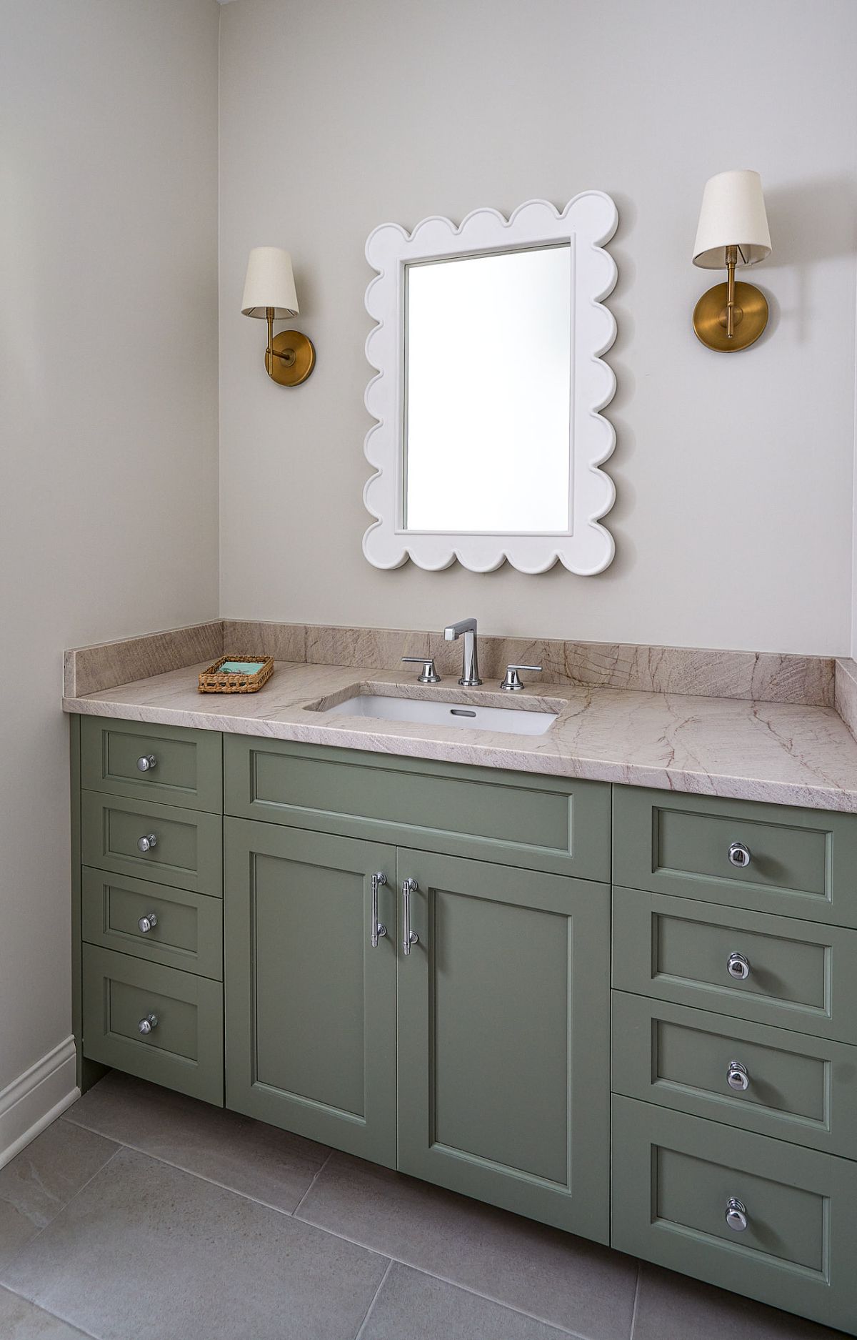 A bathroom vanity with a light green cabinet, a light-colored stone countertop, a white sink, and a white scalloped mirror, flanked by two brass wall sconces.