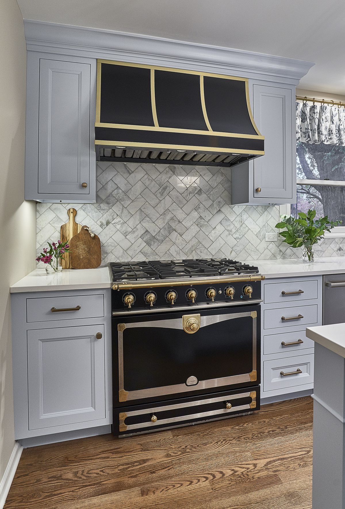 A kitchen features light gray cabinets, a black and gold range hood, a black and gold professional range, and a herringbone tile backsplash.