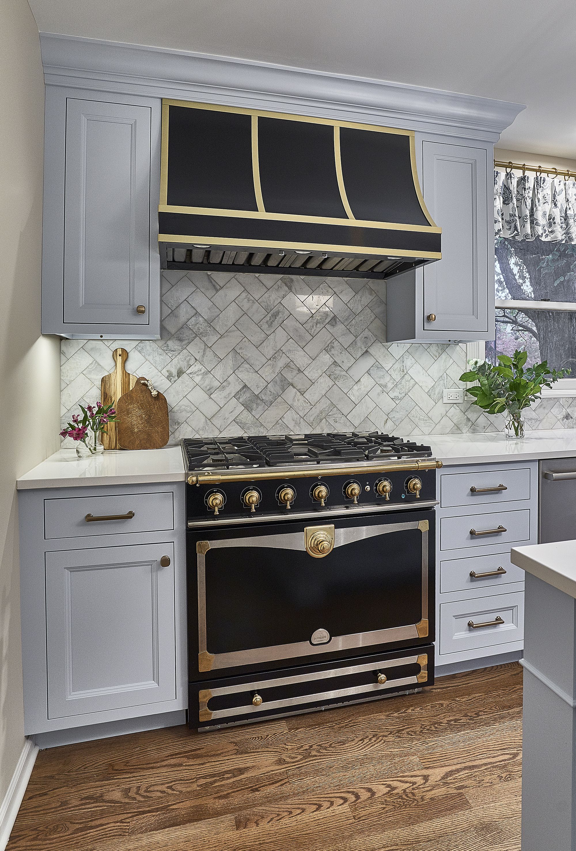 A kitchen features light gray cabinets, a black and gold range hood, a black and gold professional range, and a herringbone tile backsplash.
