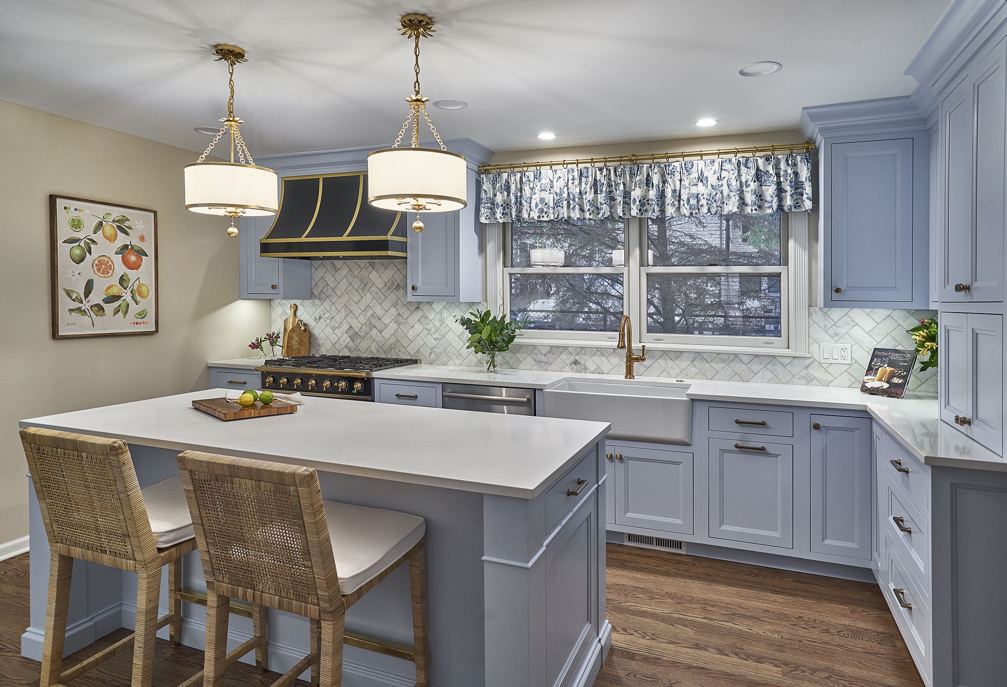 A light blue kitchen features a central island with two bar stools, a range with a black and gold hood, and a farmhouse sink under a window.