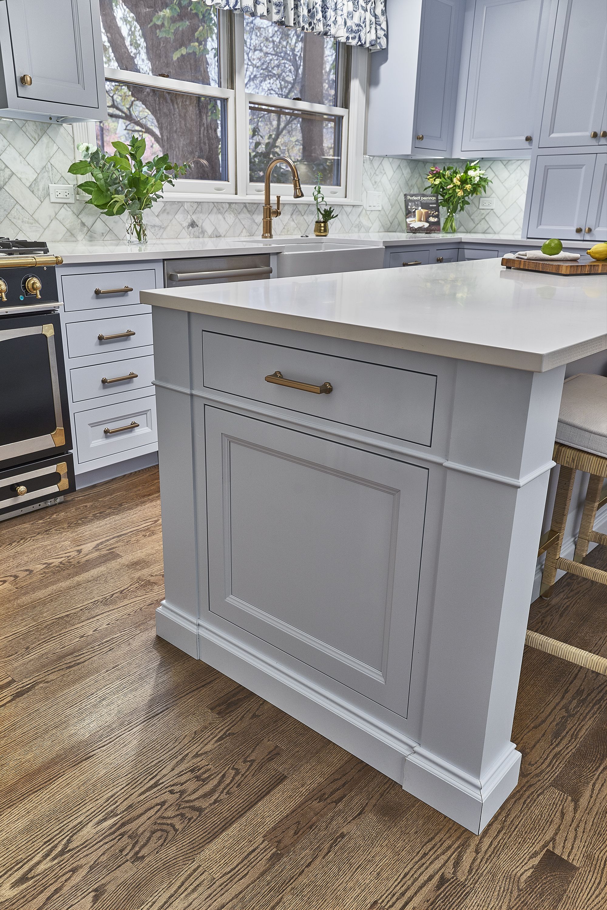 A light blue kitchen with a white countertop island, a range, and a sink under a window looking out at trees.