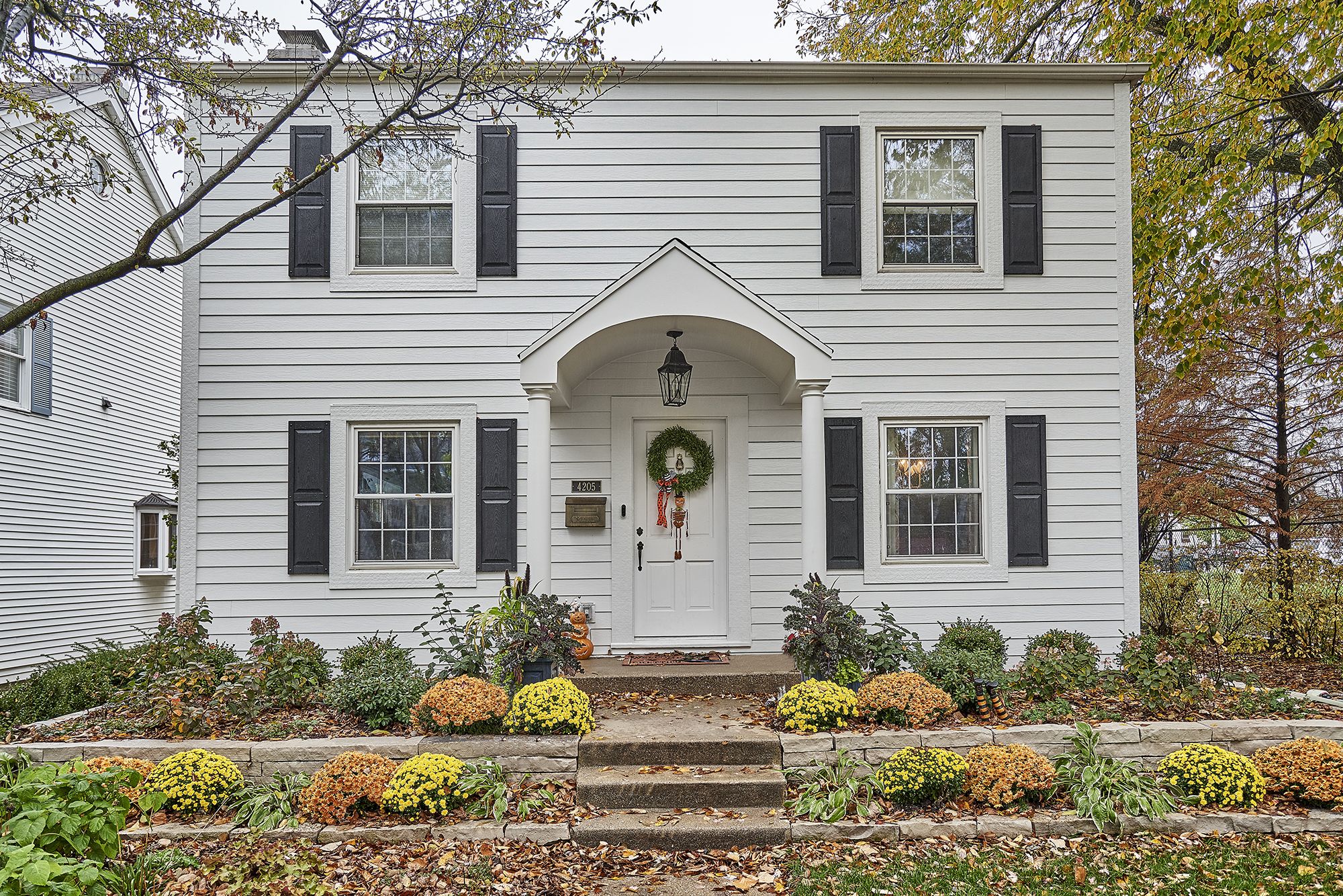 A two-story white house with black shutters, a covered front porch, and a tiered garden with colorful mums.