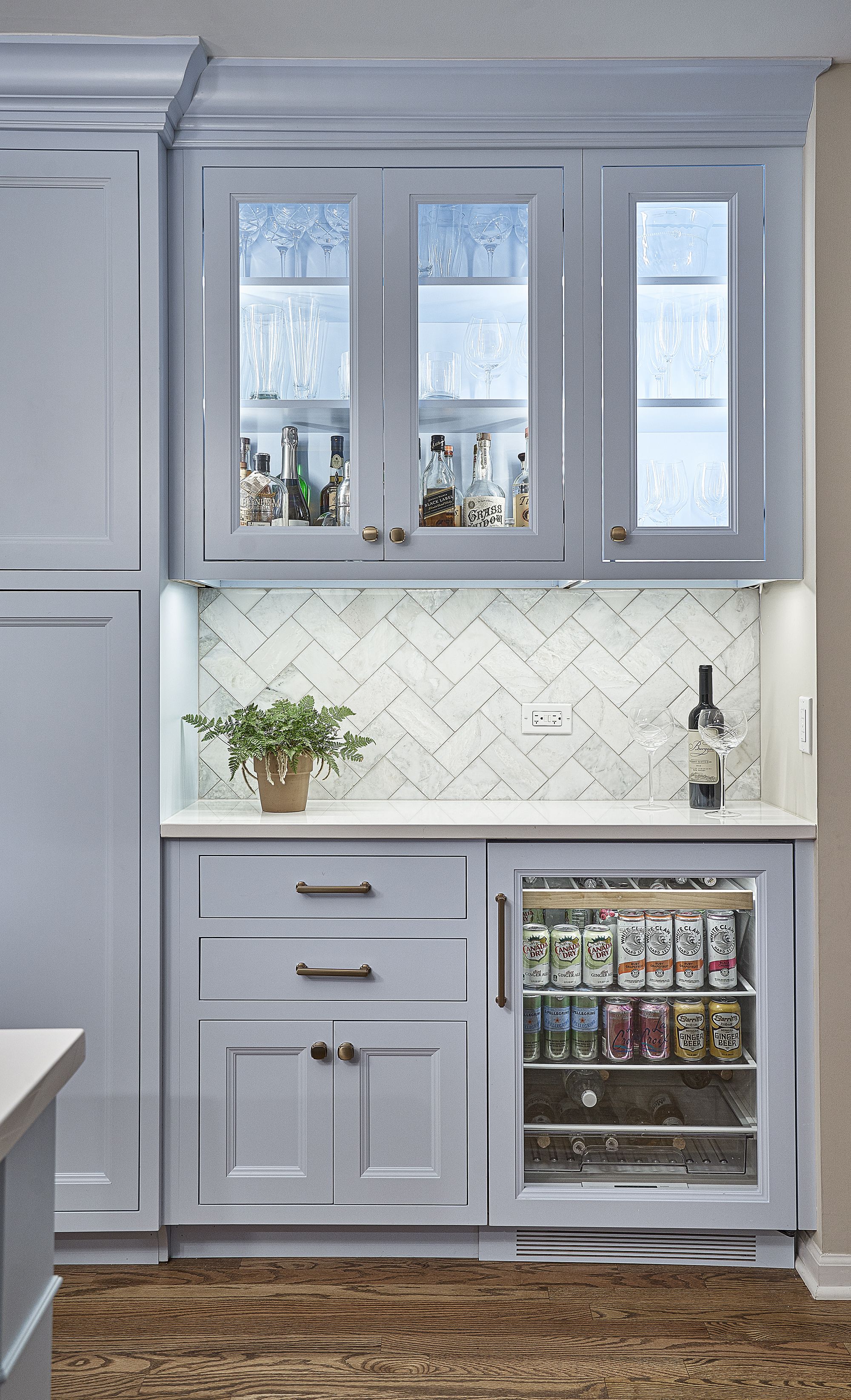 A light blue built-in bar area features glass-front upper cabinets with liquor and stemware, a white countertop, and a beverage fridge below.