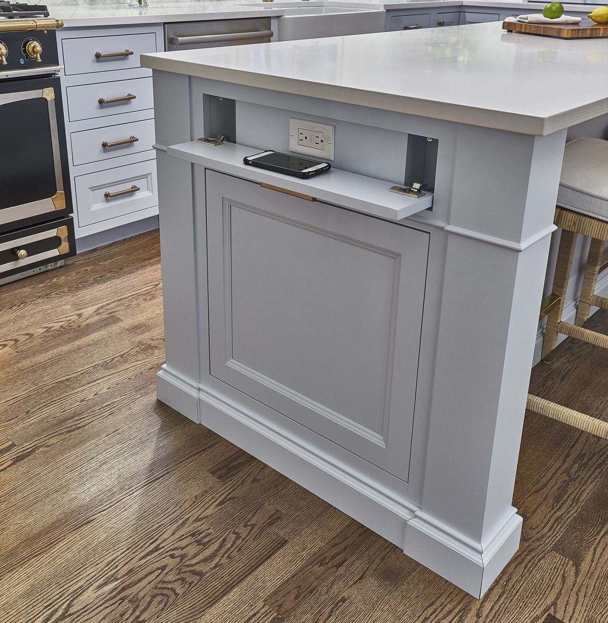 A close-up view of a kitchen island with a pull-out charging station, light gray cabinets, and dark wood flooring.