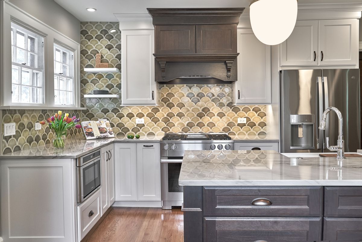 A kitchen with white upper and lower cabinets, a dark wood range hood and island, a stainless steel refrigerator, and a scallop-patterned tile backsplash.