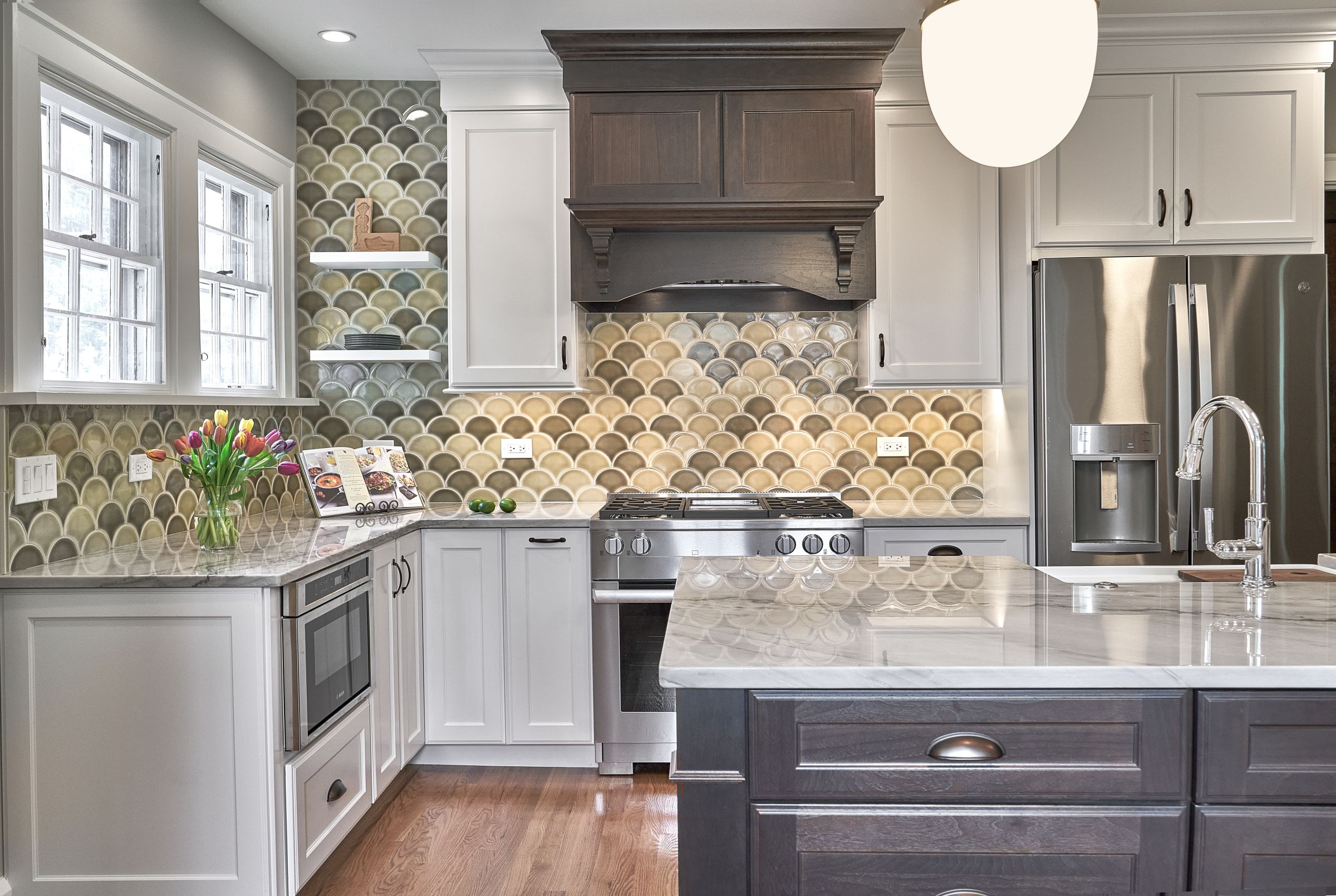 A kitchen with white upper and lower cabinets, a dark wood range hood and island, a stainless steel refrigerator, and a scallop-patterned tile backsplash.