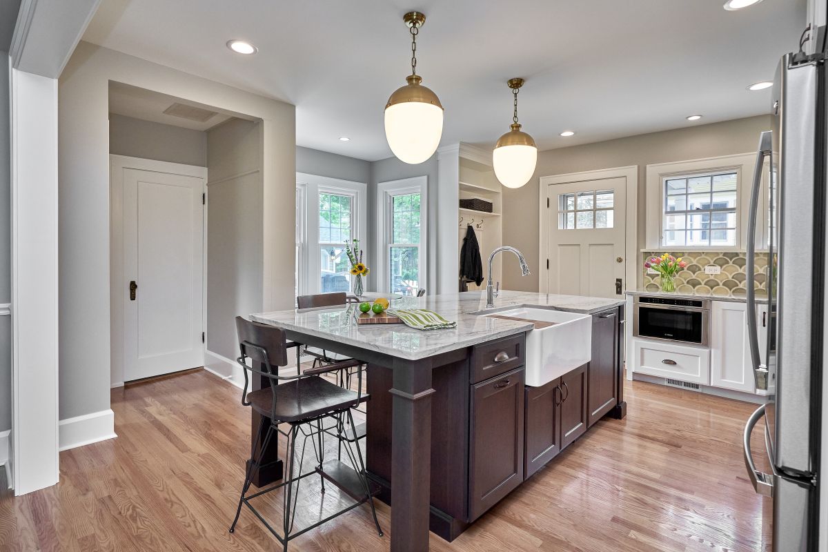 A kitchen features a dark wood island with a white farmhouse sink, light countertops, and two brass pendant lights.