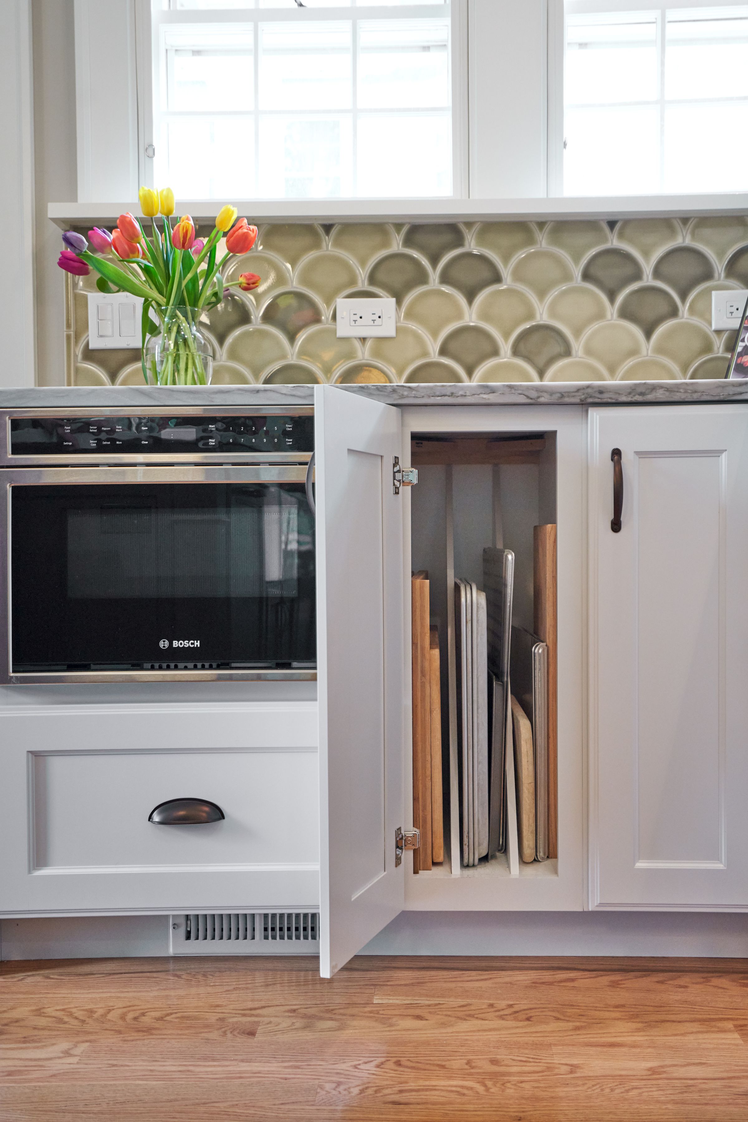 A kitchen counter with a built-in microwave, a vase of tulips, and a cabinet storing baking sheets.