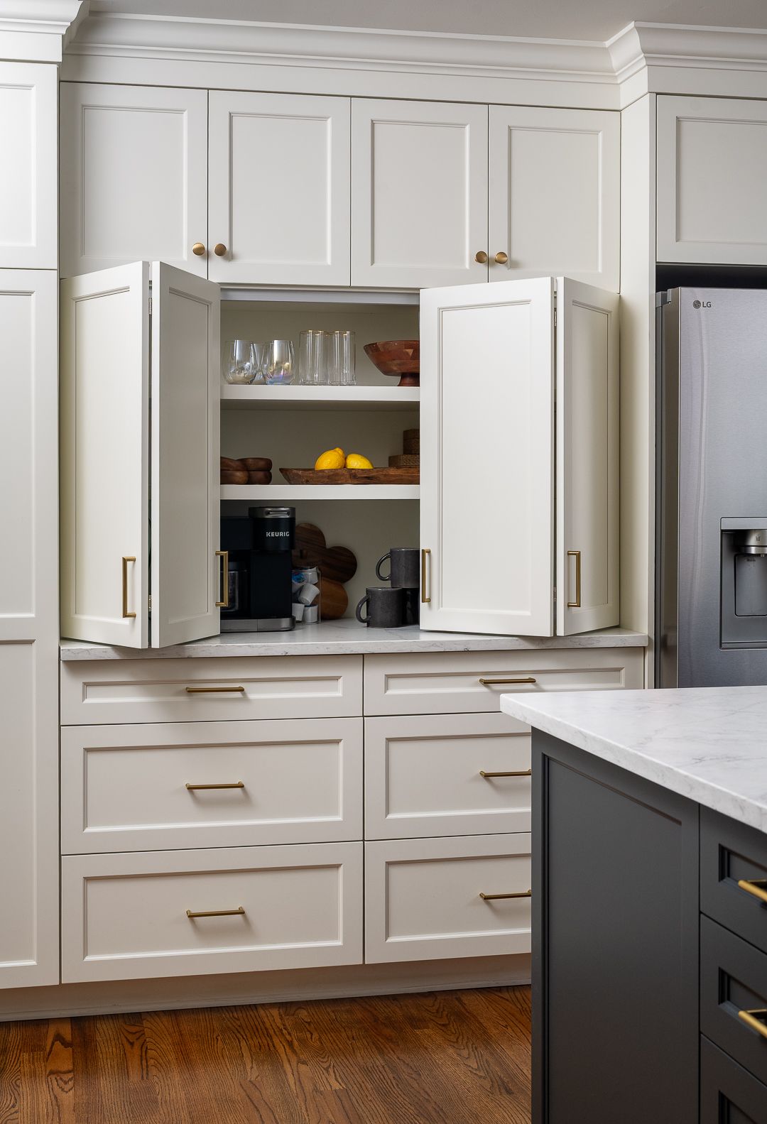 A kitchen with white upper cabinets, open pantry doors revealing shelves with dishes and a coffee maker, and white lower drawers, next to a stainless steel refrigerator and a dark gray kitchen island.