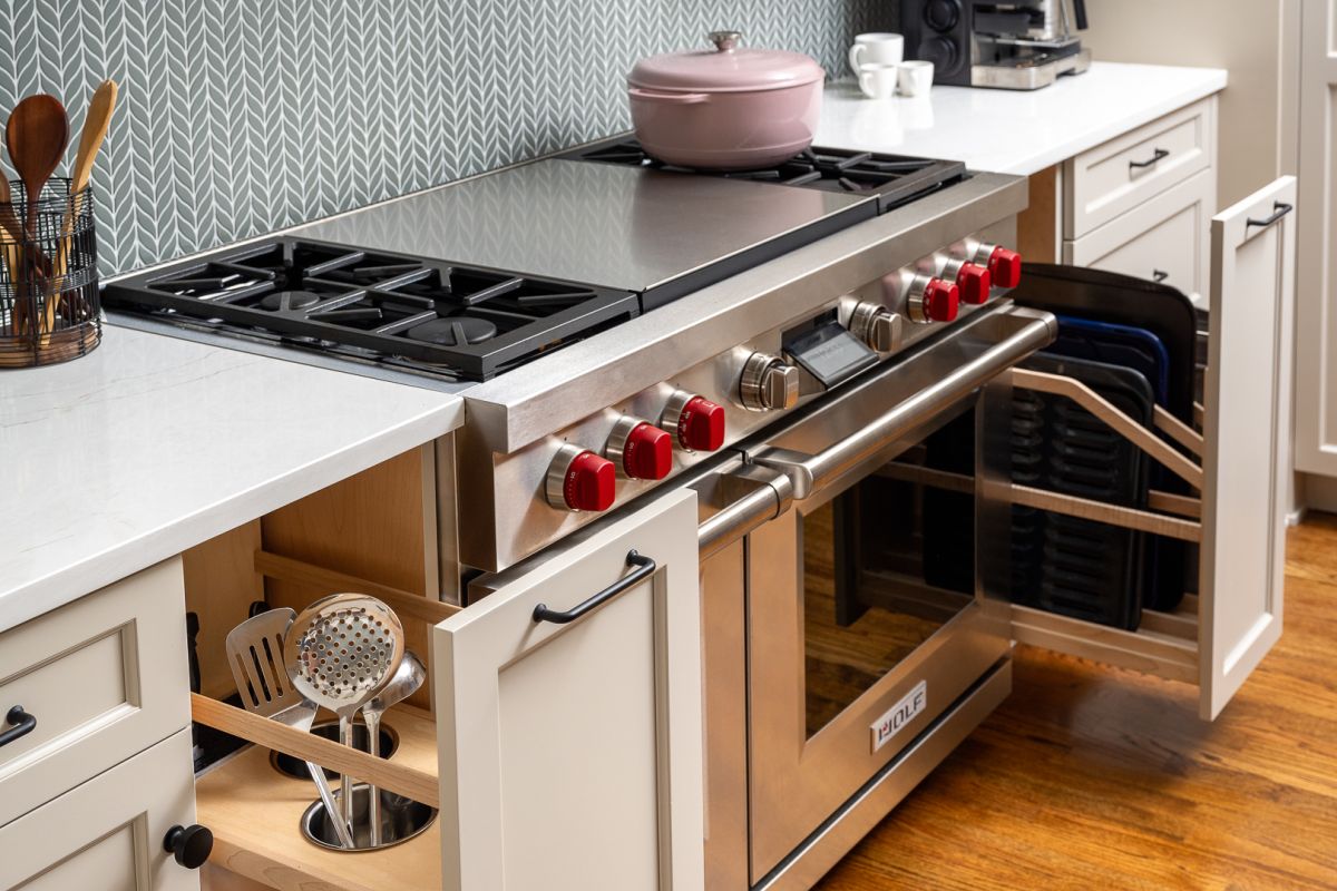 A kitchen features a stainless steel range with red knobs, white countertops, and a gray patterned backsplash. Open cabinets reveal kitchen utensils and baking sheets.