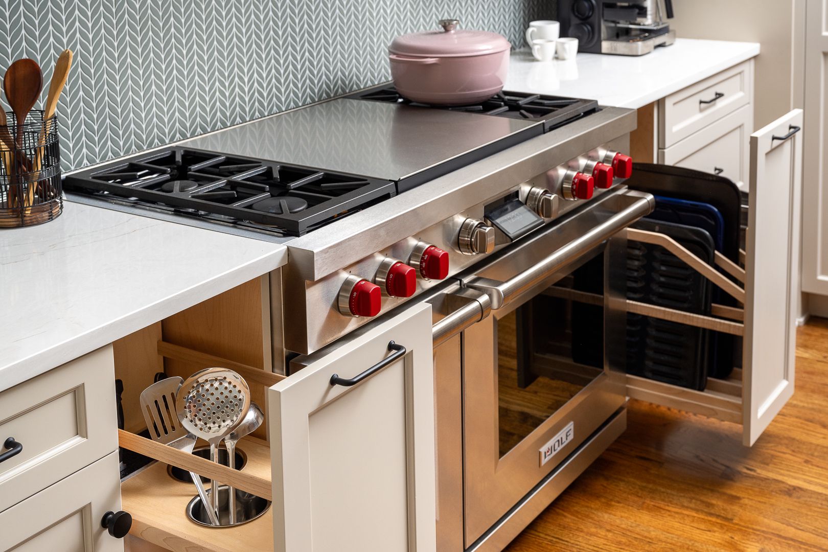 A kitchen features a stainless steel range with red knobs, white countertops, and a gray patterned backsplash. Open cabinets reveal kitchen utensils and baking sheets.