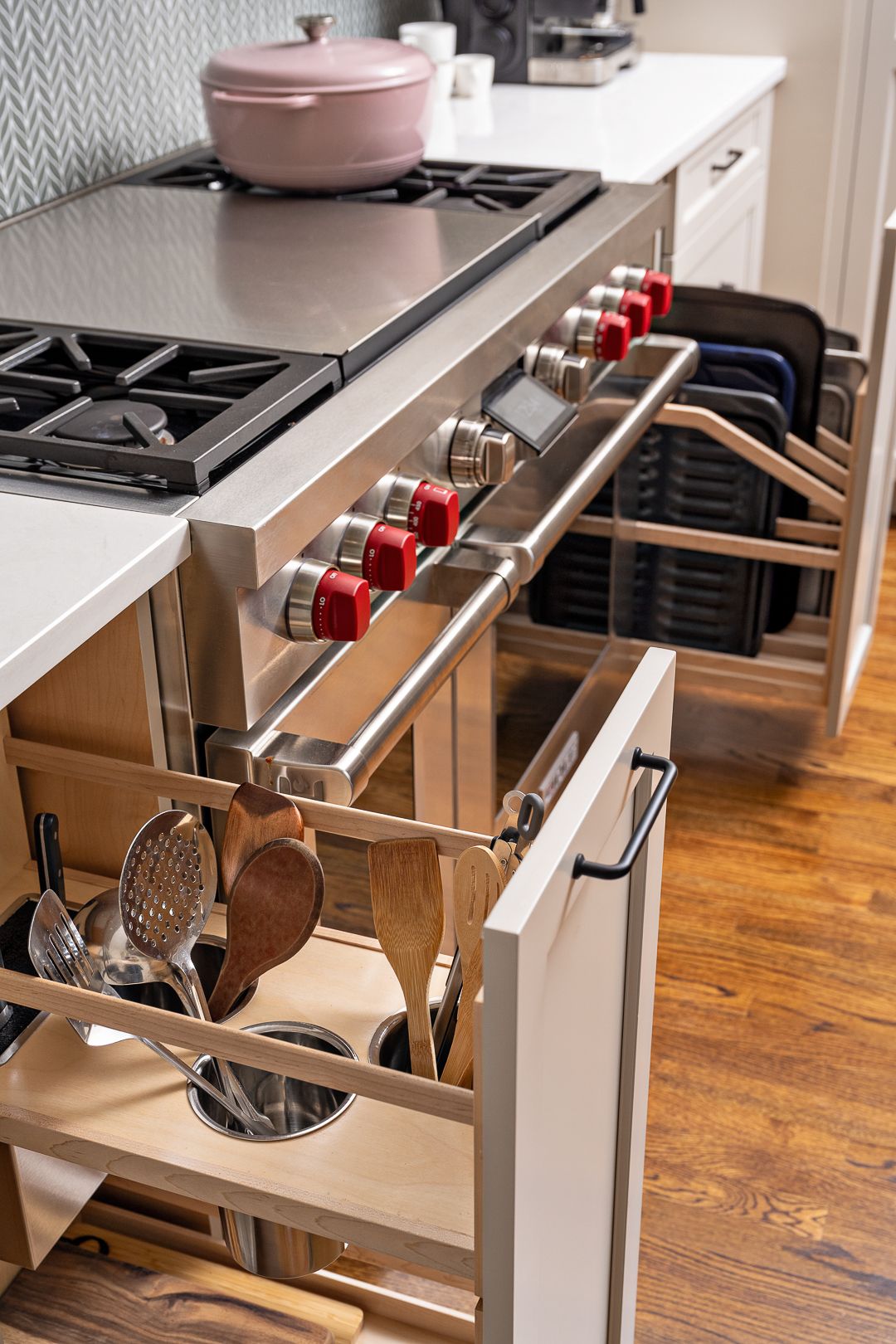 A close-up of a kitchen shows a stainless steel range with red knobs, open pull-out drawers, and a pink pot on the stovetop.