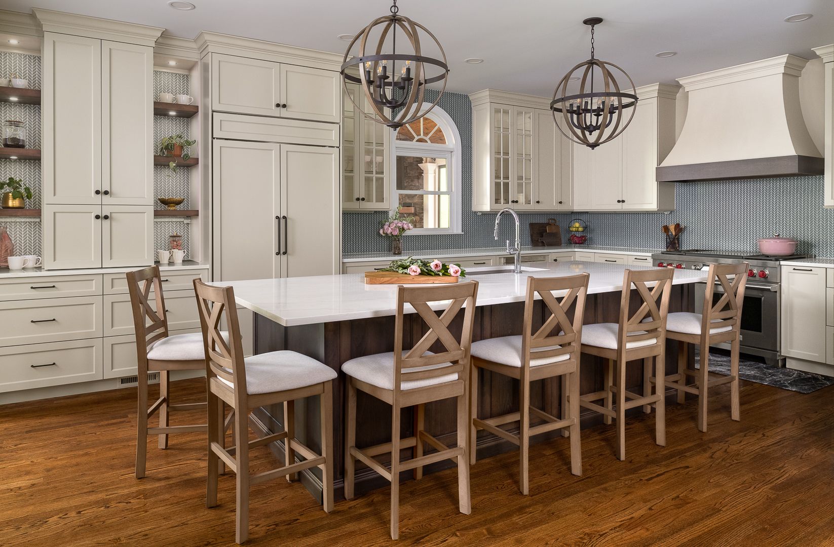 A spacious kitchen features light-colored cabinets, a large island with seating, a refrigerator, a range, and two spherical chandeliers.