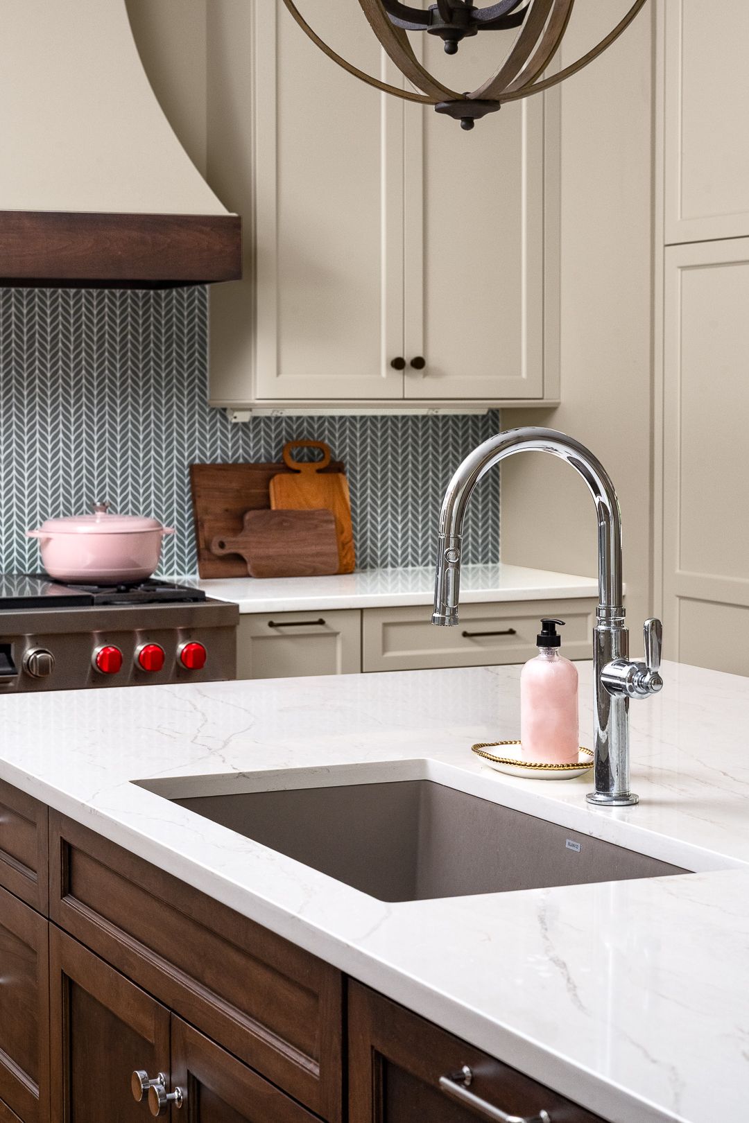 A kitchen features a white countertop island with a dark sink and chrome faucet, dark wood lower cabinets, and light upper cabinets with a herringbone tile backsplash.
