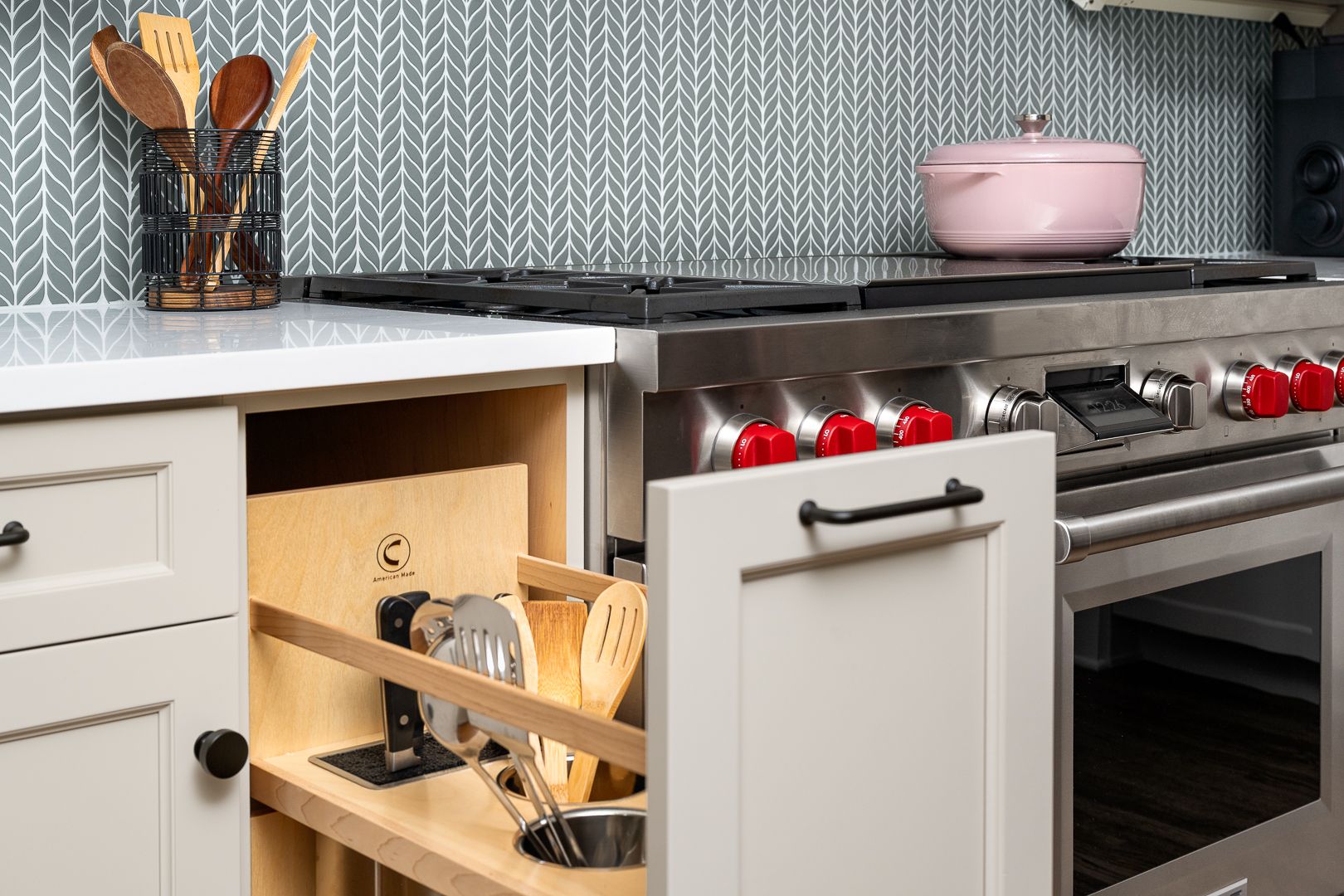 A kitchen with a stainless steel range, light gray cabinets, and a gray and white patterned backsplash. An open cabinet drawer reveals kitchen utensils.