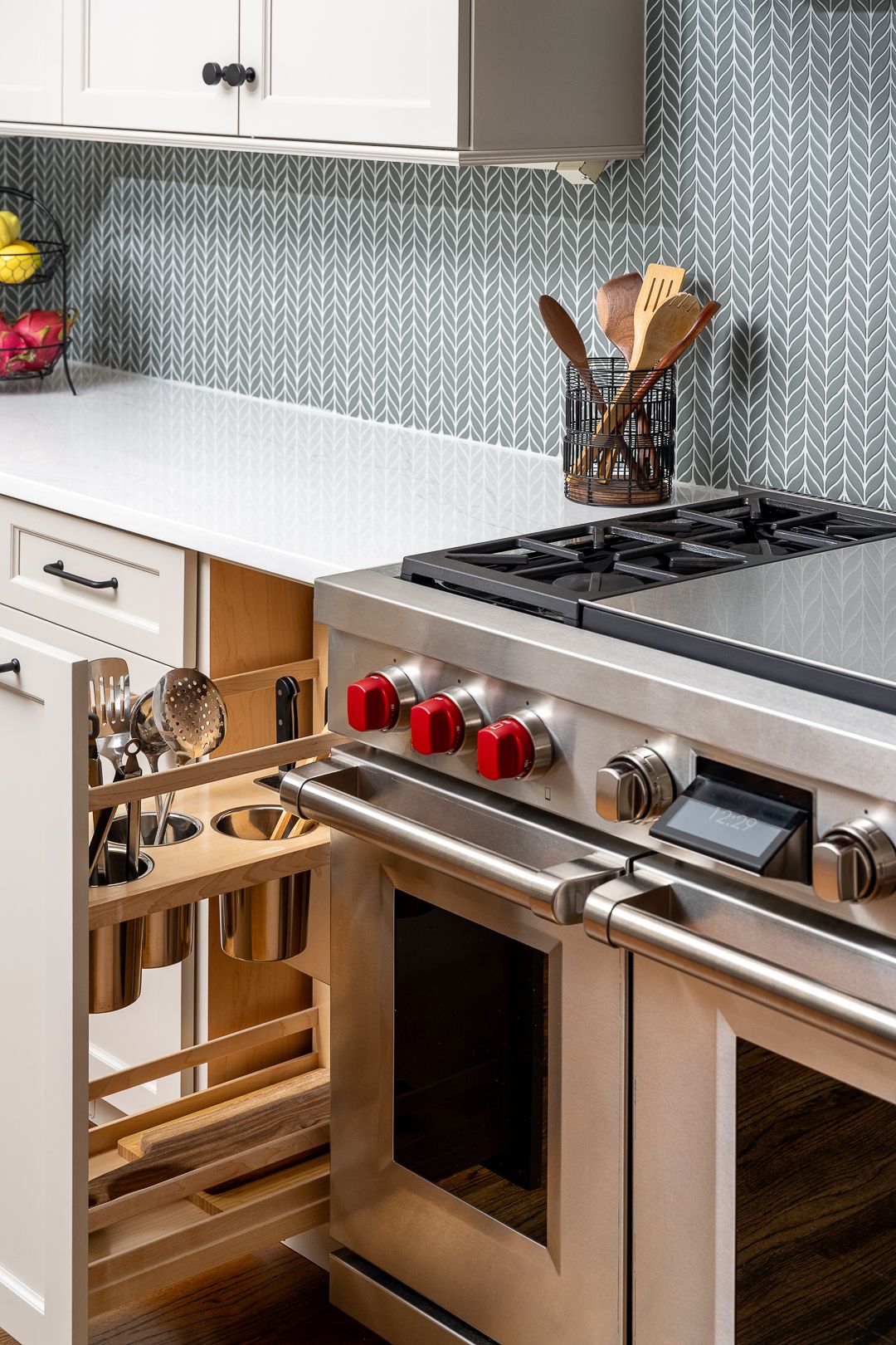A kitchen features white cabinets, a light-colored countertop, a gray and white patterned backsplash, and a stainless steel range with red knobs. A pull-out cabinet next to the range holds kitchen utensils.