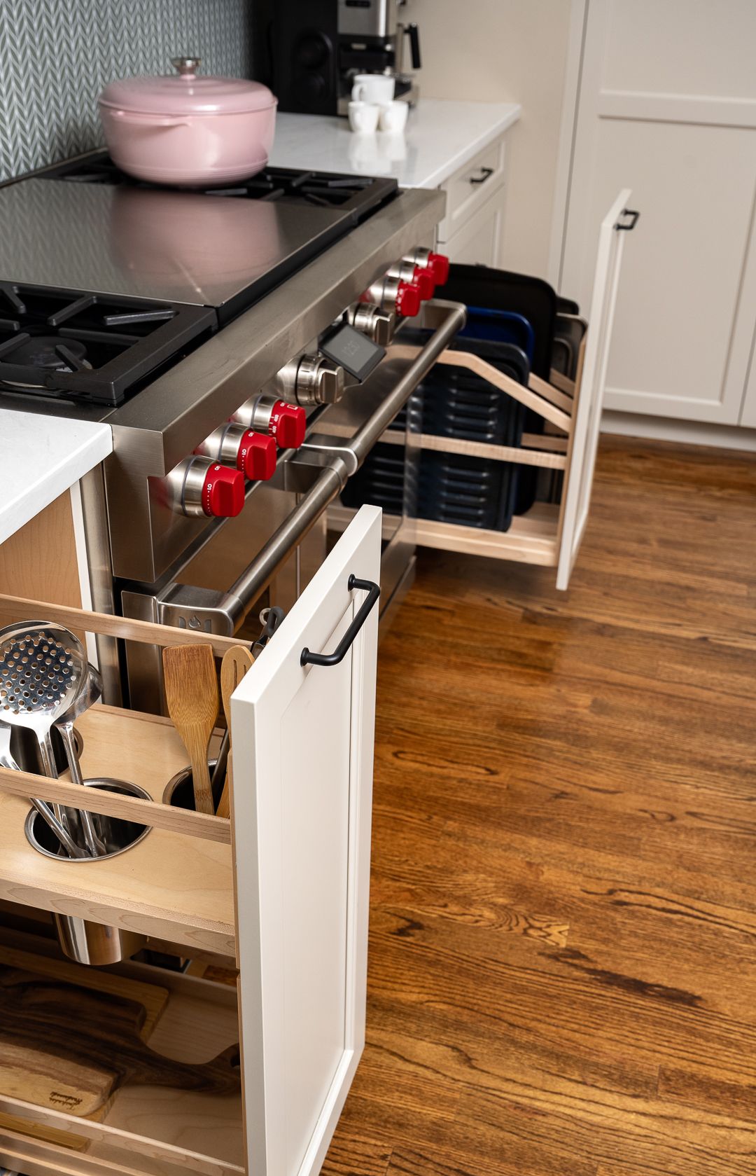 A kitchen features a stainless steel range with red knobs, white cabinets with pull-out storage, and hardwood floors.