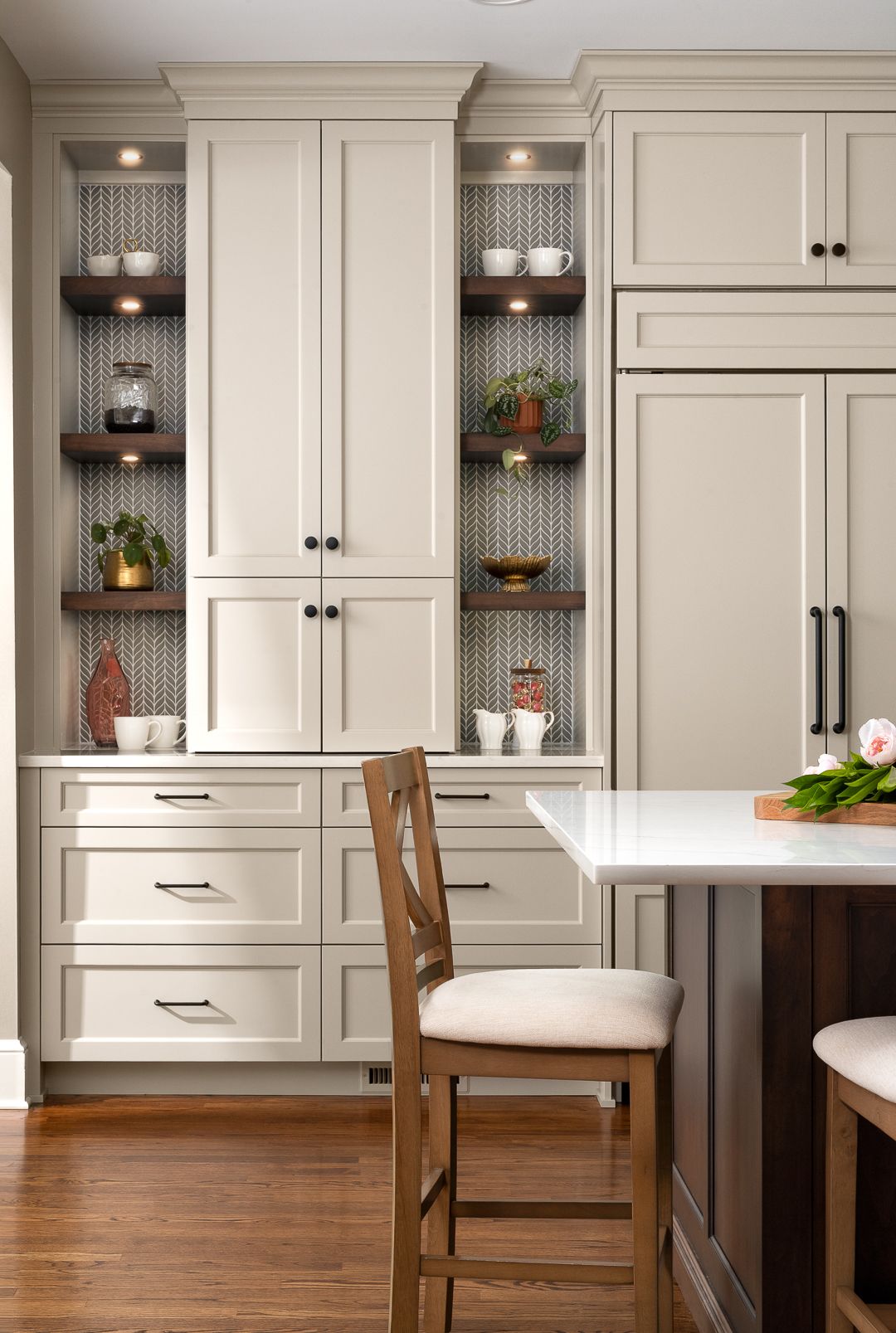 A kitchen features light-colored cabinetry, open shelving with decorative items, a built-in refrigerator, and a kitchen island with bar stools.
