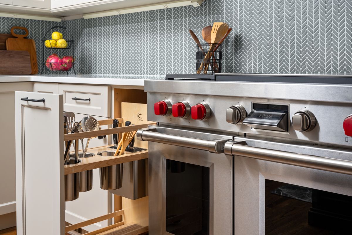 A kitchen with white cabinets, a gray herringbone tile backsplash, and a stainless steel range with red knobs. A pull-out cabinet next to the range holds kitchen utensils.