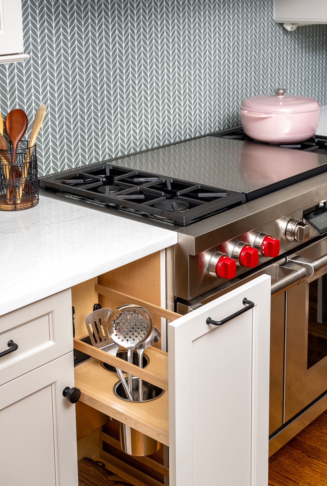 A kitchen features a stainless steel range with a griddle, white countertops, and a gray patterned backsplash. A pull-out cabinet holds cooking utensils.