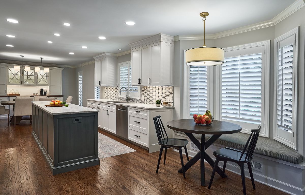 A kitchen features white upper and lower cabinets, a dark gray island, a dining table with chairs, and a window seat.