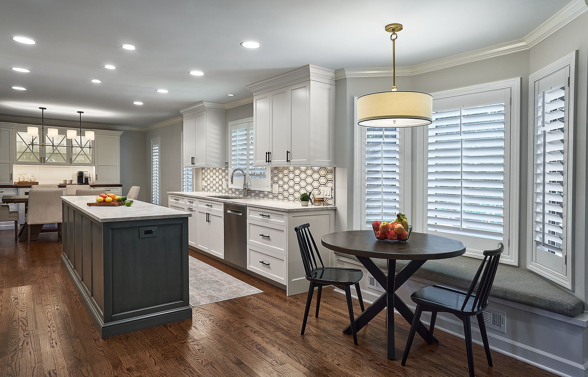 A kitchen features white upper and lower cabinets, a dark gray island, a dining table with chairs, and a window seat.