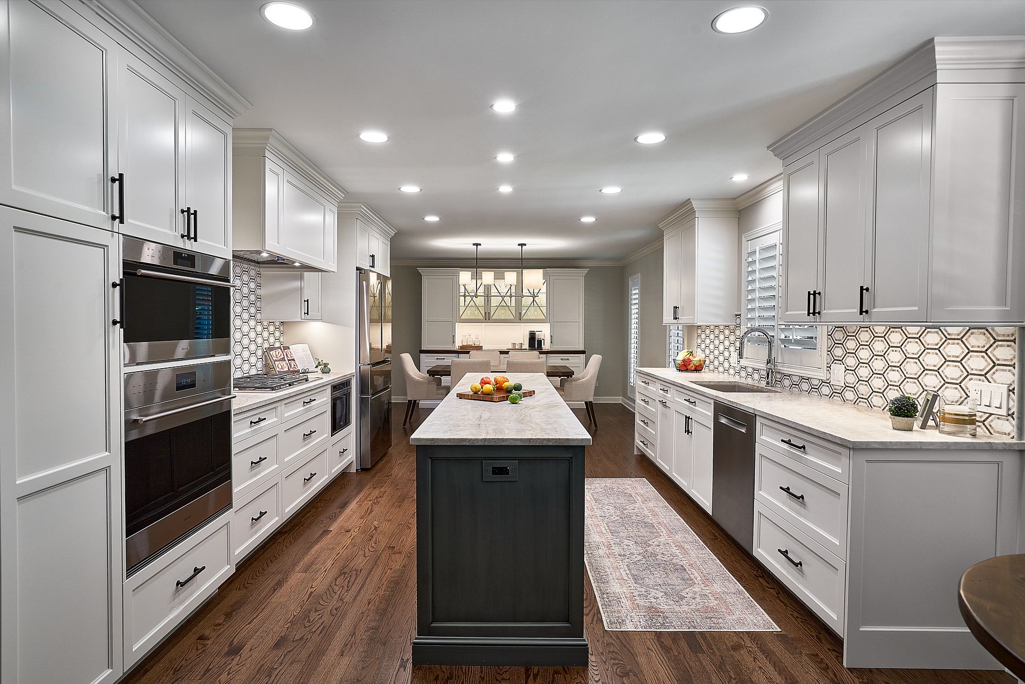 A long, bright kitchen features white shaker cabinets, dark wood floors, a dark gray island, and a dining area beyond.