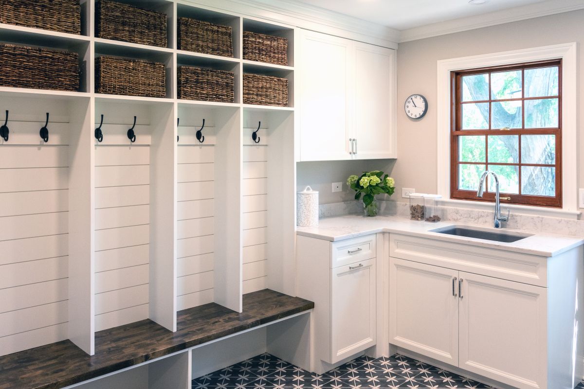 A mudroom and laundry area with white built-in cubbies, dark wood benches, white cabinets, a sink, and a window.