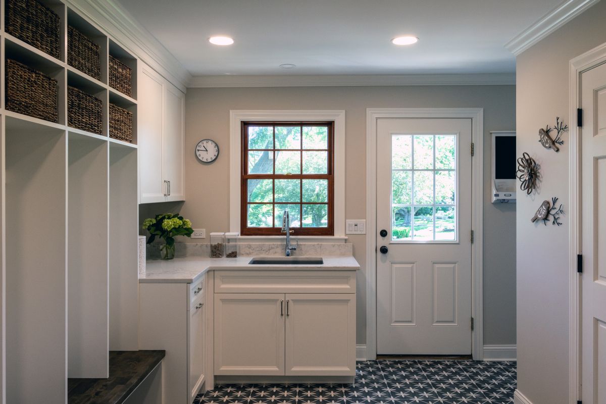 A mudroom and laundry area with white built-in shelving, a sink under a window, a patterned floor, and a white door leading outside.