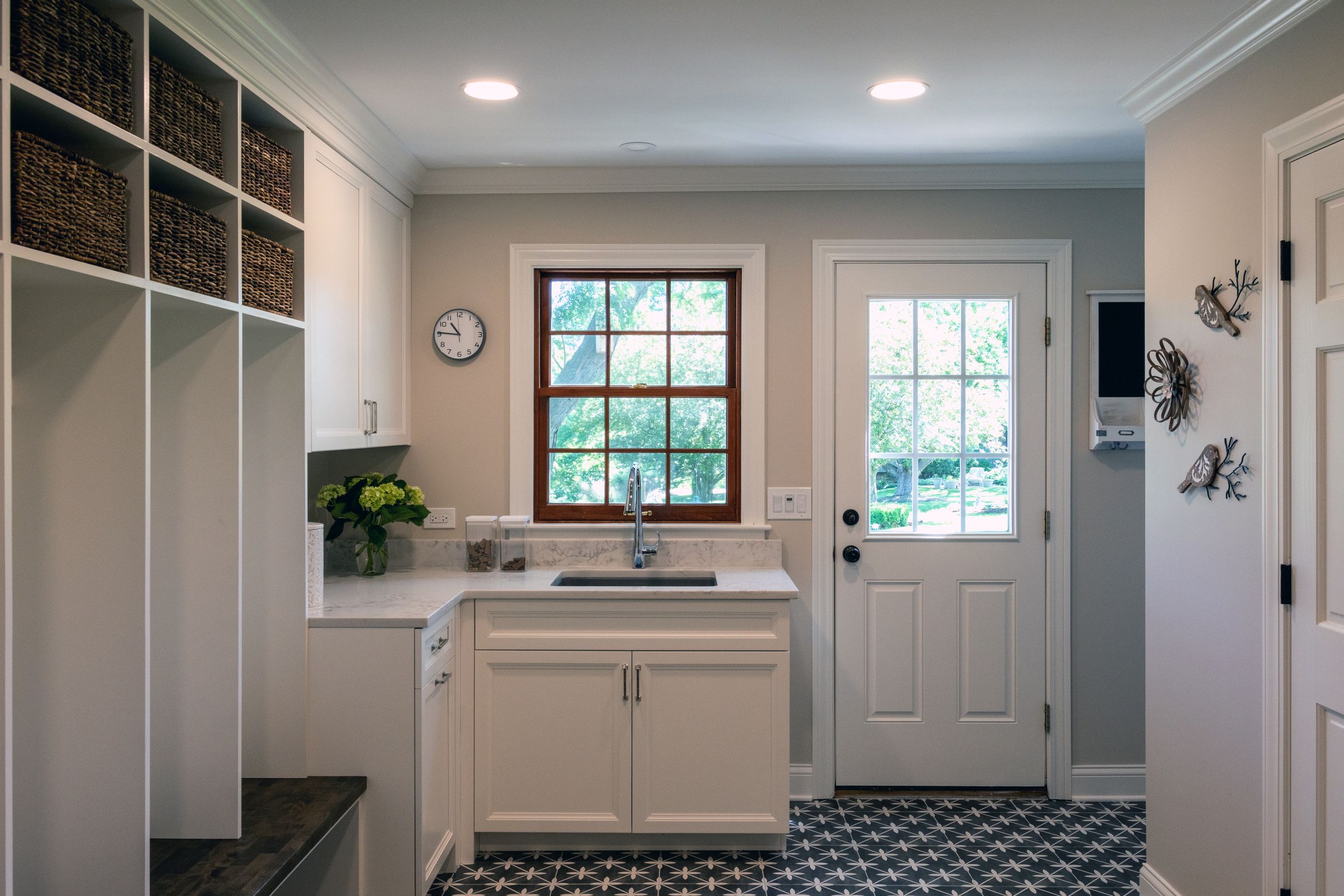 A mudroom and laundry area with white built-in shelving, a sink under a window, a patterned floor, and a white door leading outside.