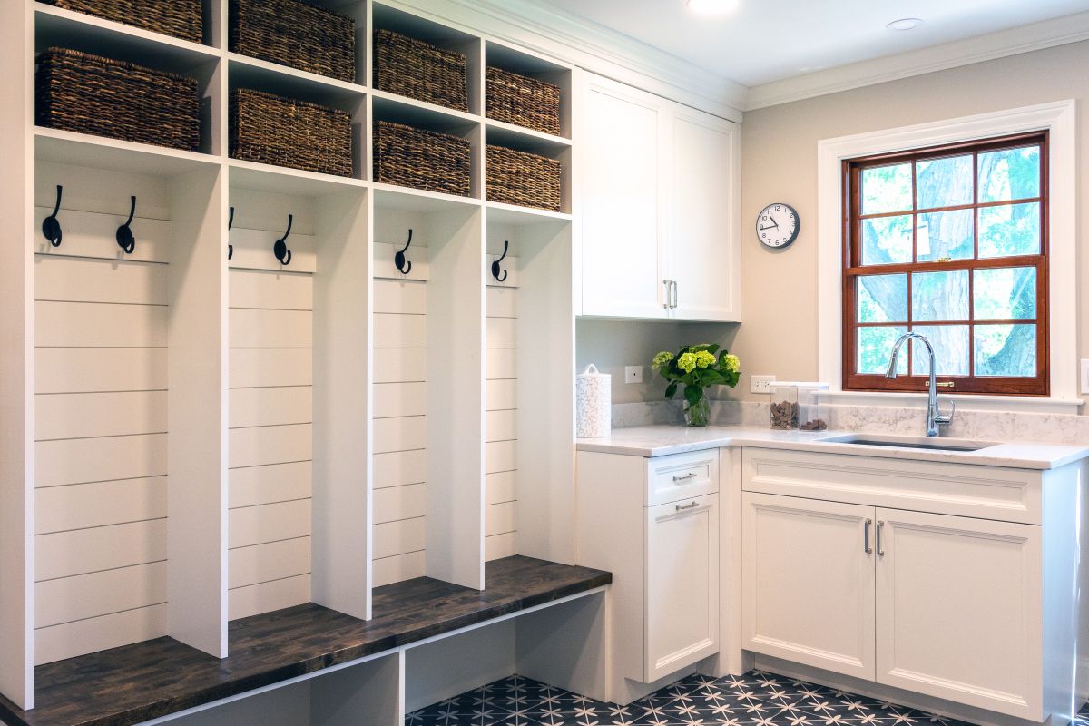 A mudroom and laundry area features white built-in cubbies with wicker baskets, a dark wood bench, and a white sink cabinet with a window.