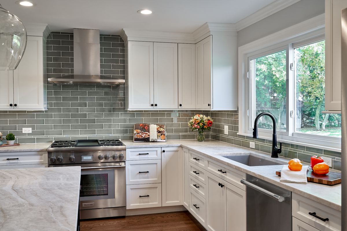 A white kitchen features a stainless steel range, hood, and appliances, with gray subway tile backsplash and light countertops.