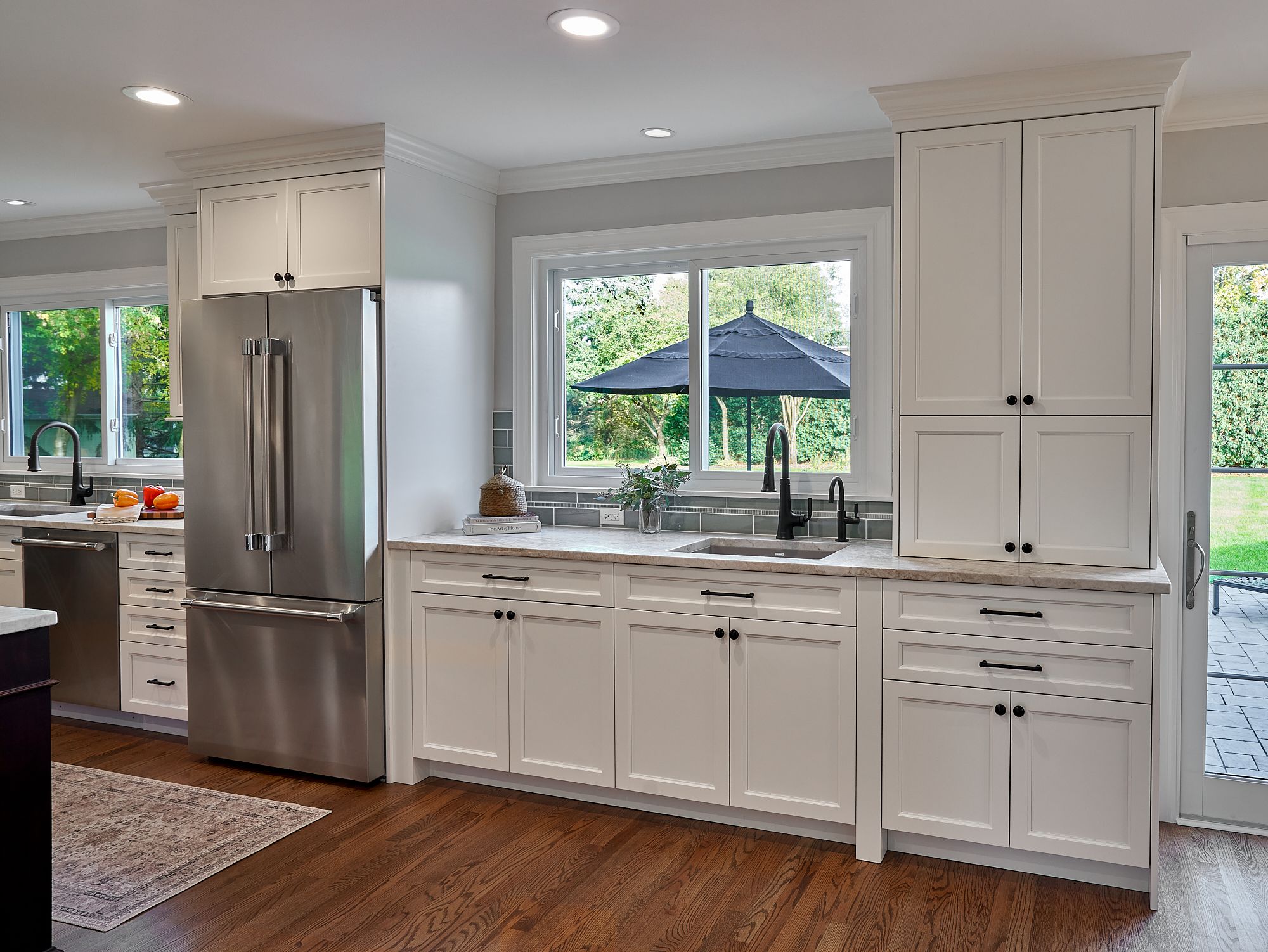 A kitchen with white shaker cabinets, stainless steel appliances, dark wood floors, and a view of a backyard through large windows.