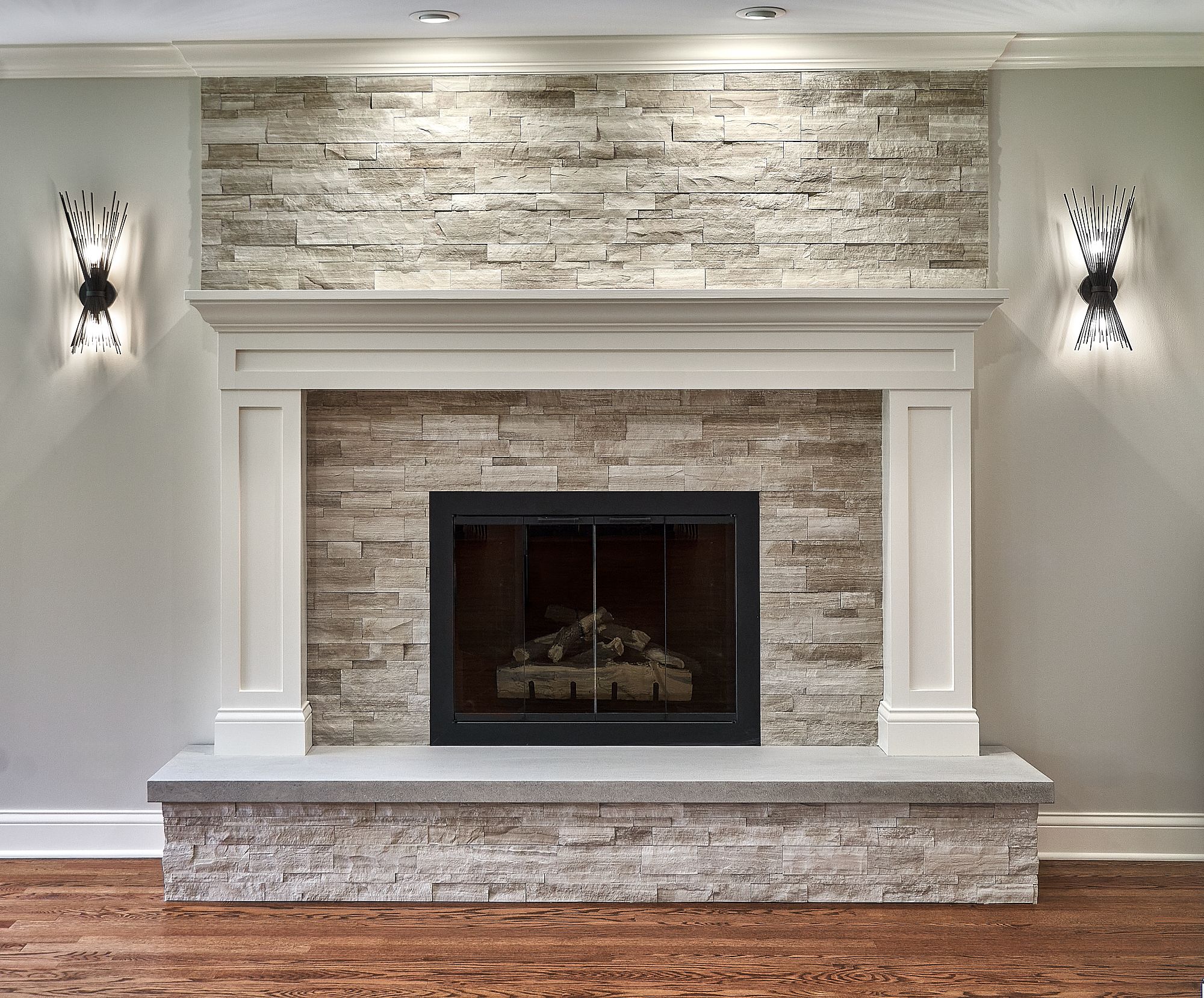A fireplace with a light-colored stacked stone surround and hearth, a white mantel, and two sconces on the light gray walls.