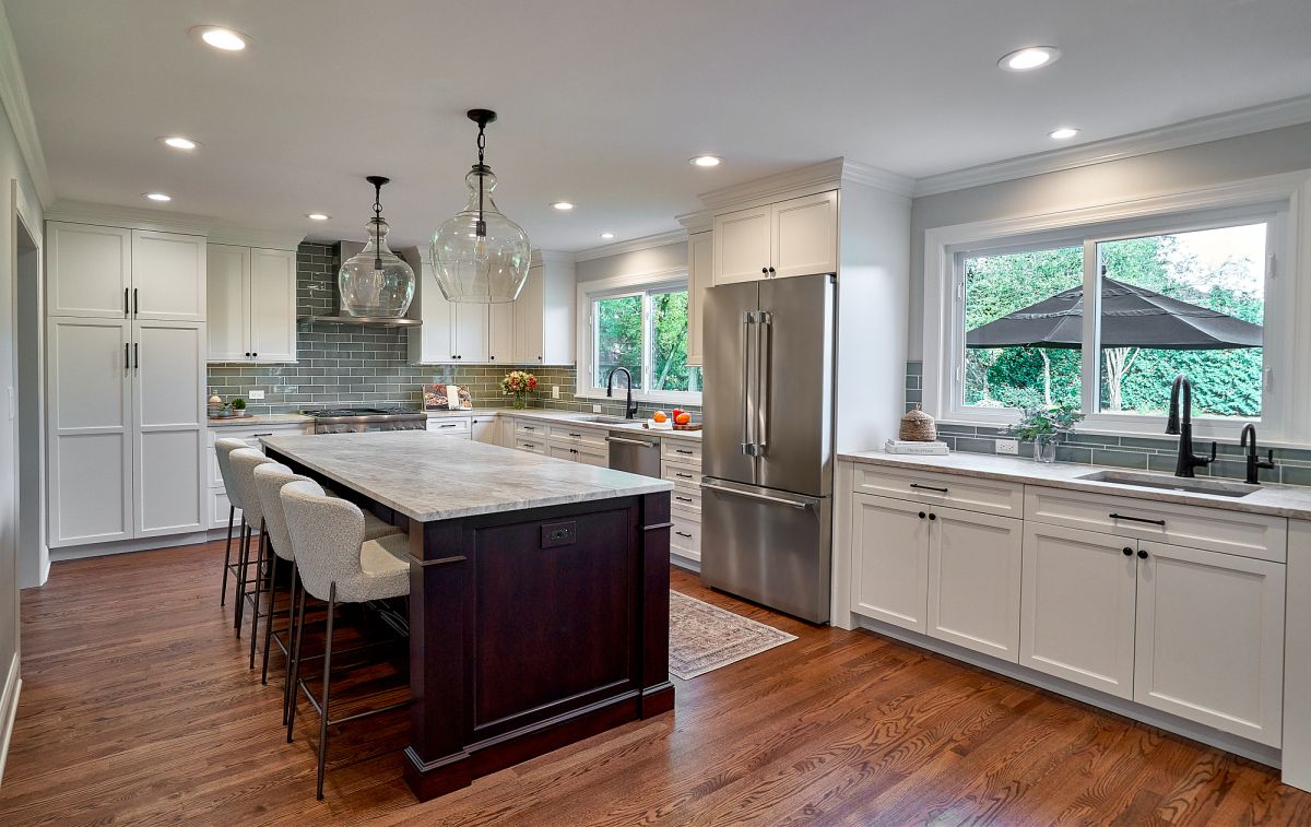 A bright kitchen features white shaker cabinets, a large dark wood island with a light stone top, and stainless steel appliances.