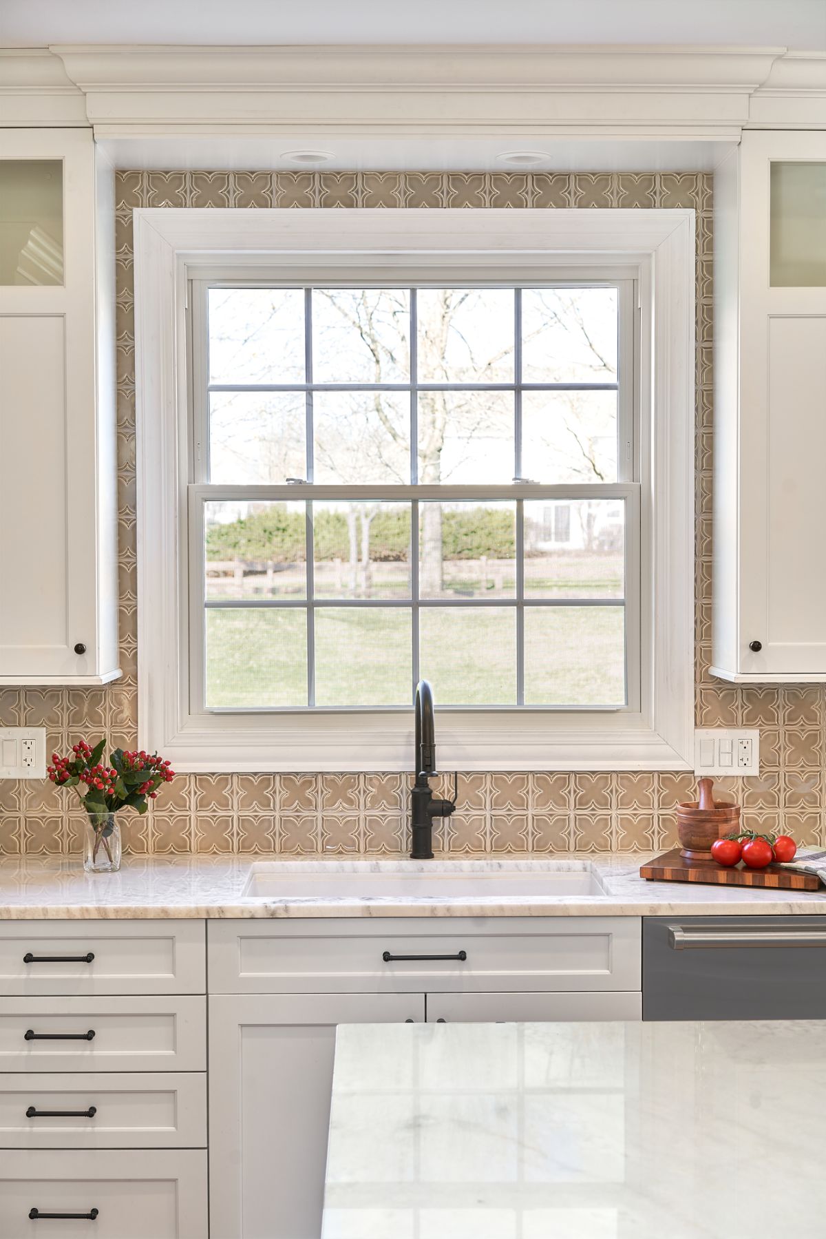 A kitchen features a large window above a sink, surrounded by patterned tile backsplash and white cabinetry.