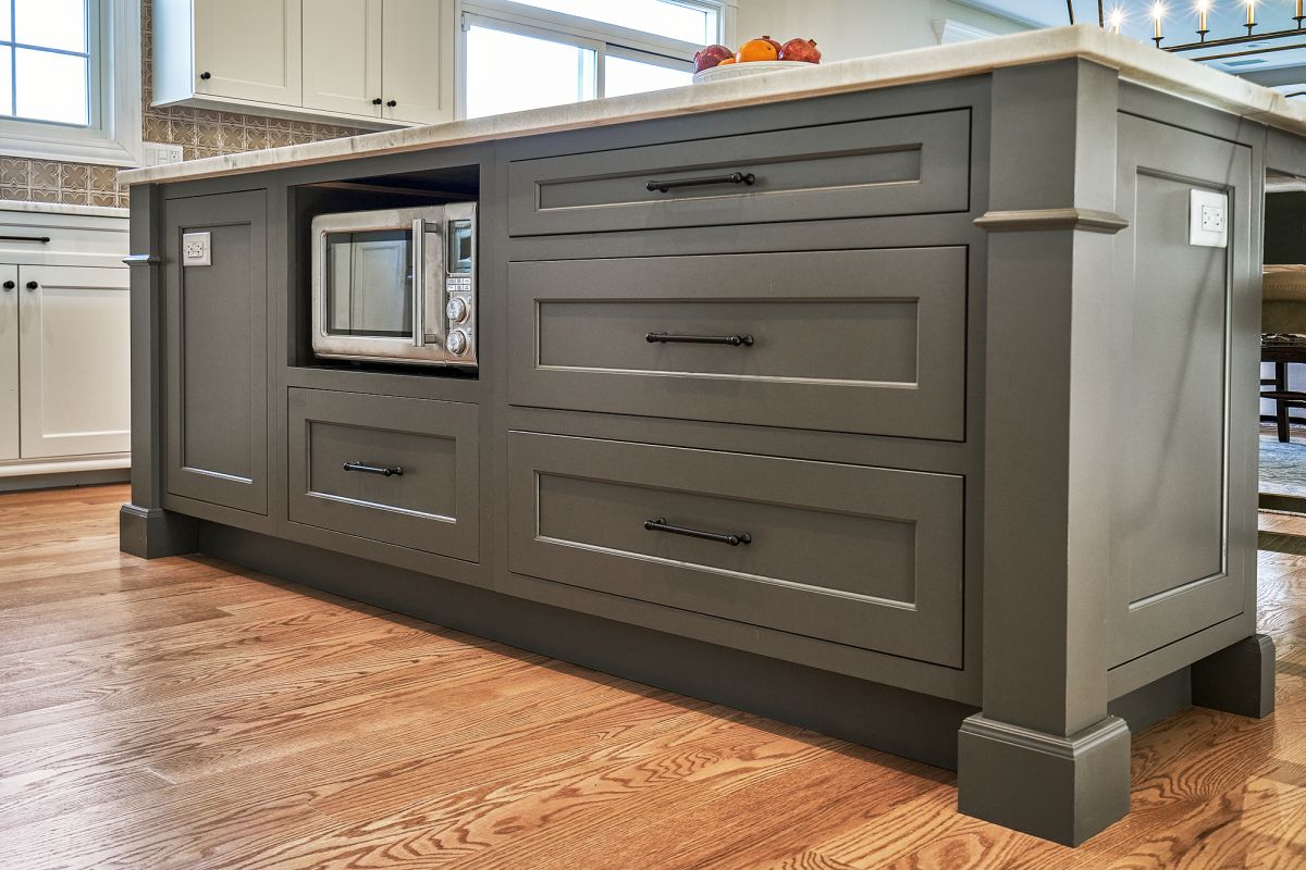 A kitchen island with dark gray shaker-style cabinets, a built-in microwave, and a light-colored countertop. Hardwood floors are visible in the foreground.