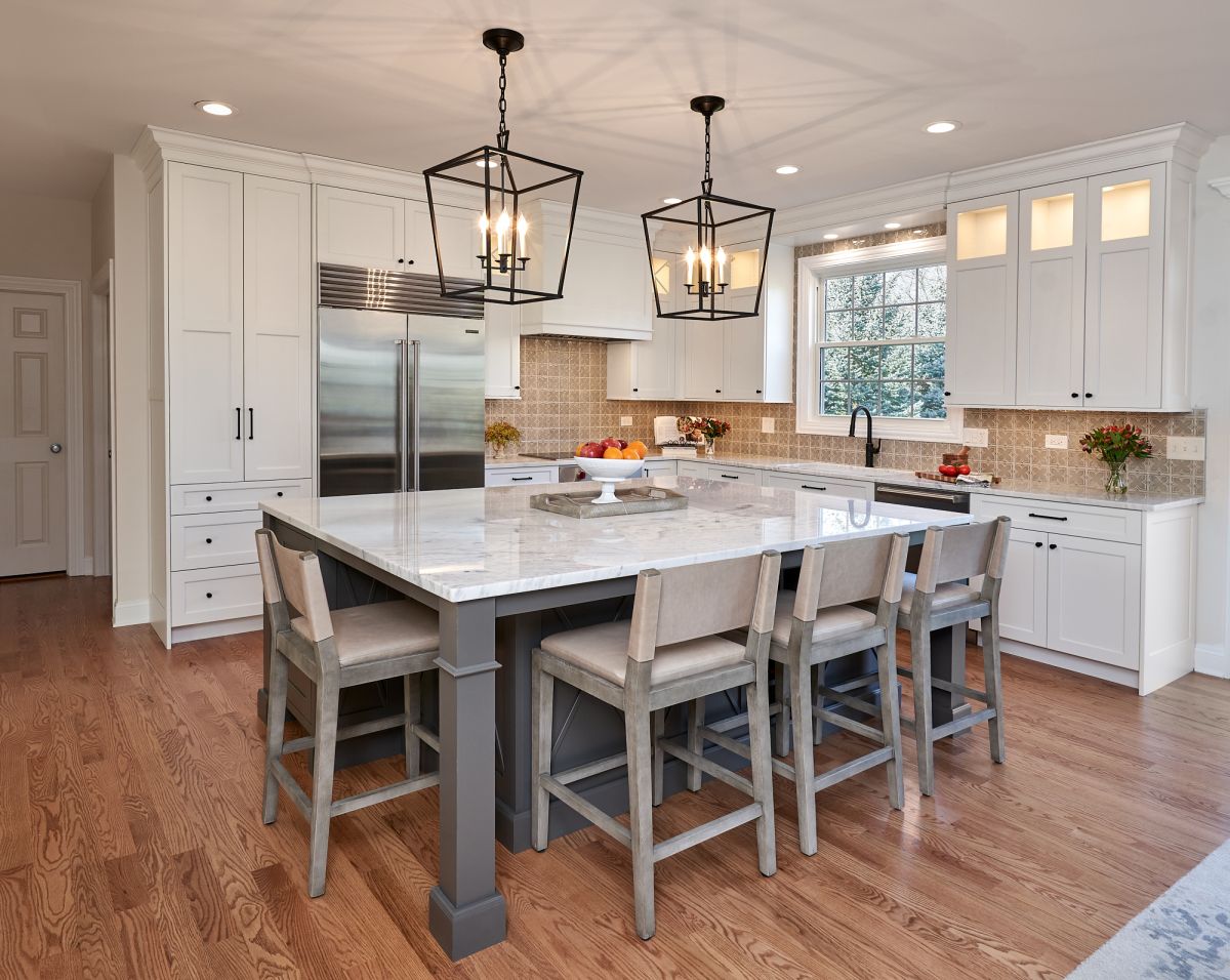 A white kitchen features a large gray island with four bar stools, white cabinetry, stainless steel appliances, and wood floors.