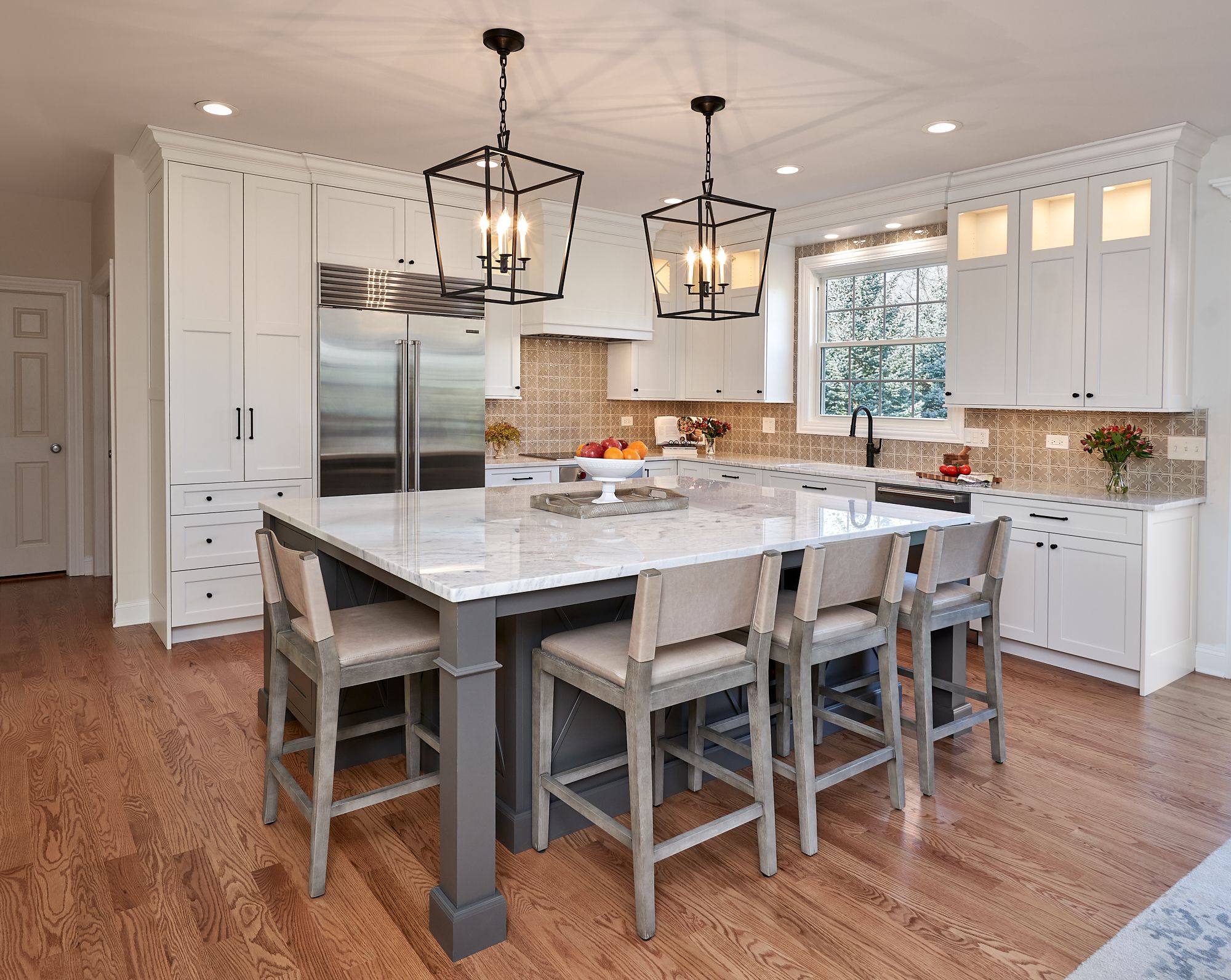 A white kitchen features a large gray island with four bar stools, white cabinetry, stainless steel appliances, and wood floors.