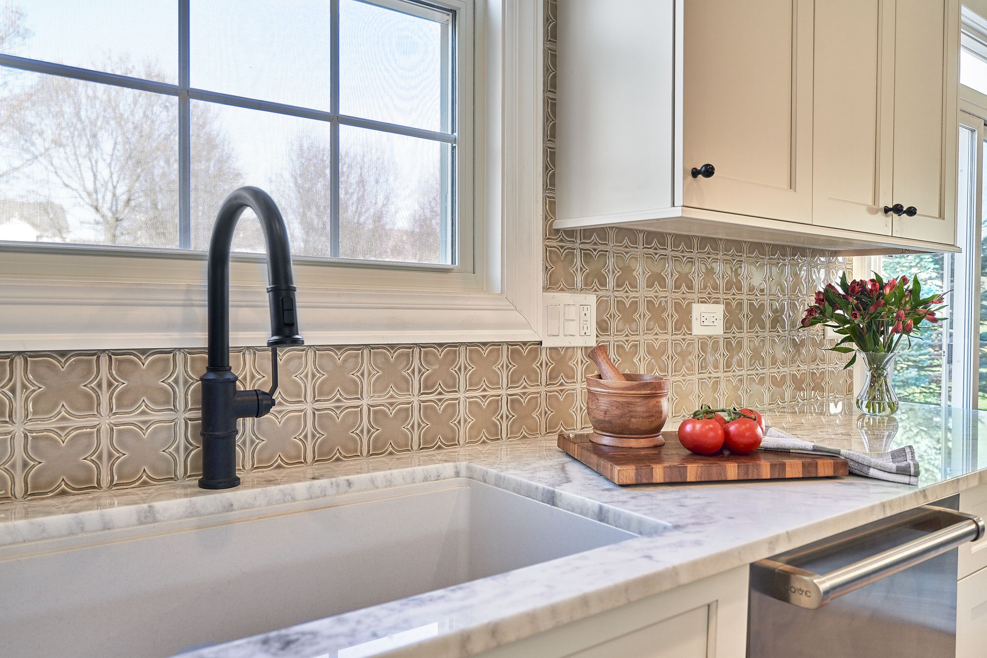 A close-up of a kitchen sink area with a black faucet, patterned backsplash, and light-colored cabinets.