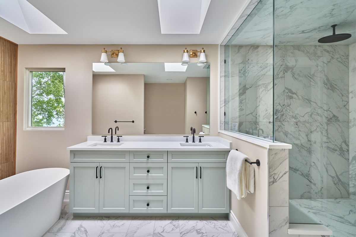 A primary bathroom with a light green double vanity, a freestanding white bathtub, a walk-in shower, and two skylights.